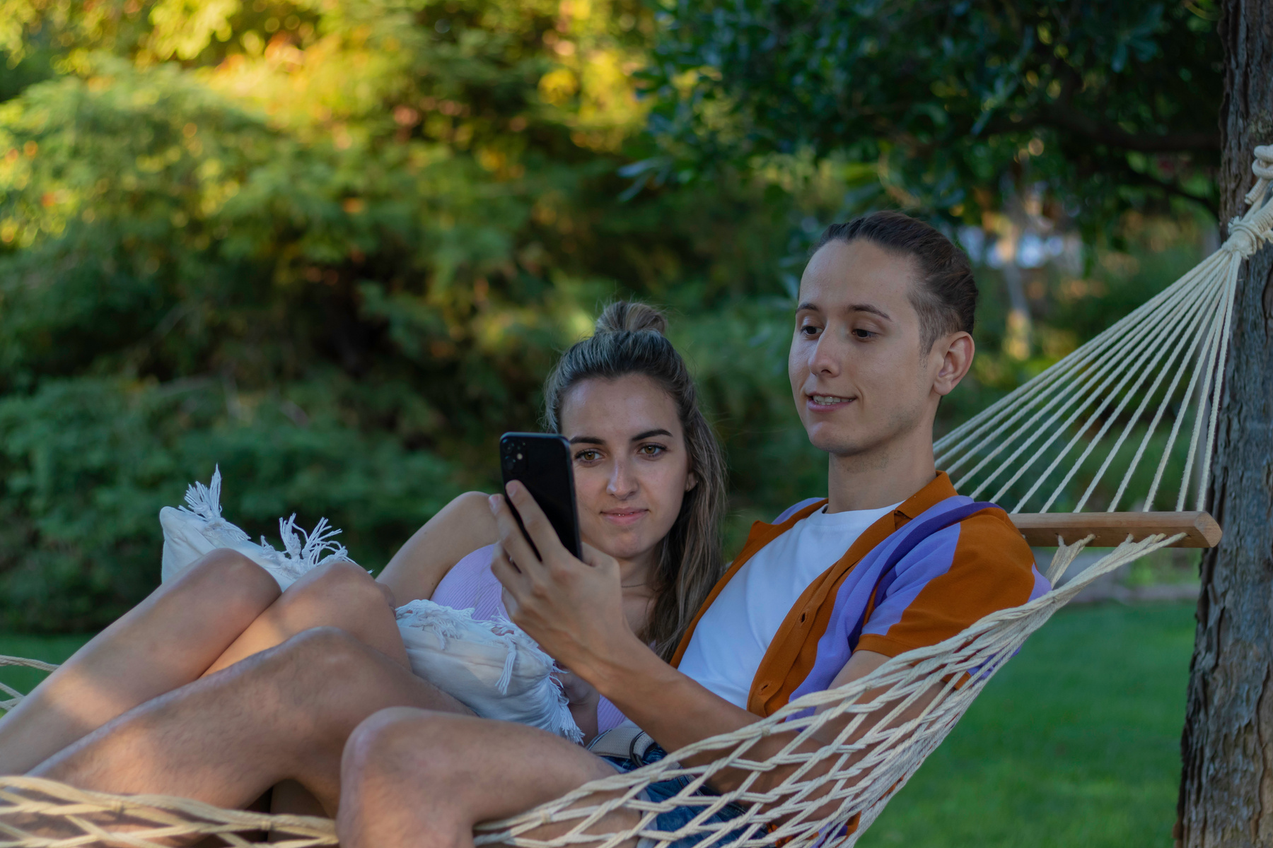 Young modern couple sitting in a hammock in holidays and vactions while looking to the smartphone sorrounded by nature